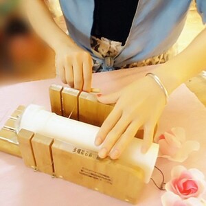 May include: A person using a wooden soap cutter to cut a white bar of soap. The soap cutter has a metal wire and a wooden handle. The soap is on a pink surface.