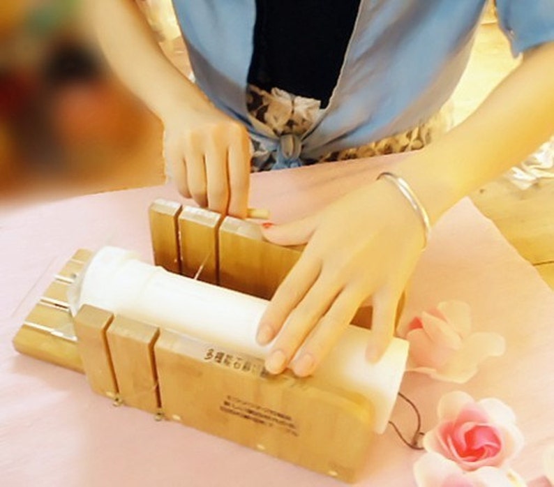 May include: A person using a wooden soap cutter to cut a white bar of soap. The soap cutter has a metal wire and a wooden handle. The soap is on a pink surface.