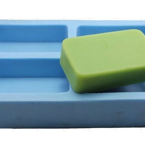 Puede incluir: Un molde de silicona azul con cuatro cavidades rectangulares para hacer barras de jabón. Una barra de jabón verde está parcialmente insertada en una de las cavidades.