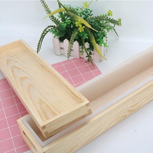 May include: A wooden soap mold with a rectangular shape and a removable lid. The mold is made of light-colored wood and has a white interior. The lid is placed next to the mold. A small white picket fence with greenery is in the background.