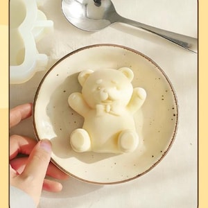 May include: A light-colored bear-shaped confection with a bow tie, presented on a small, off-white plate with a speckled rim. A stainless steel spoon rests nearby. A hand gently holds the plate, suggesting a culinary or decorative theme.