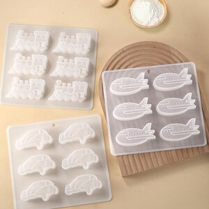 May include: Three silicone molds for baking. The molds are white and feature different designs: a train, an airplane, and a car. Each mold has six cavities.