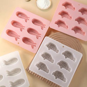 May include: Two silicone baking molds, one pink and one white, with six cavities each. The pink mold features airplane shapes, while the white mold features car shapes.