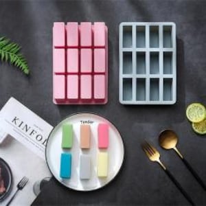 Peut inclure: Deux moules rectangulaires en silicone, un rose et un gris, chacun doté de douze cavités rectangulaires. Une assiette blanche présente six petits blocs rectangulaires colorés en vert, pêche, rose, bleu, gris et jaune, avec le mot Tender imprimé.