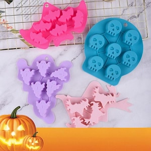 May include: Four silicone molds in pink, blue, purple, and light pink, featuring cavities shaped like bats, skulls, ghosts, and witches. The molds are arranged on a wire rack and a marble surface, accompanied by two orange jack-o'-lantern pumpkins.