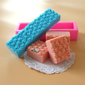 May include: A collection of handmade soaps and molds. The soaps are peach-colored and feature a rose pattern. A blue soap mold is filled with blue soap, also with a rose pattern. A pink rectangular mold is in the background. The soaps are on a white doily.