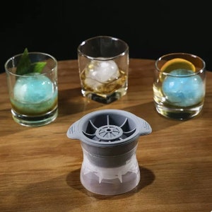 Könnte beinhalten: Eine graue Silikon-Eisform mit Kugelform. Die Form befindet sich auf einer Holzoberfläche mit drei Gläsern, die mit Eiskugeln gefüllt sind. Die Eiskugeln sind blau, klar und weiß.
