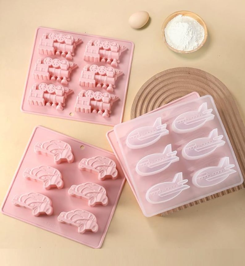 May include: Two pink silicone baking molds. One mold has six airplane shapes and the other has six train shapes.