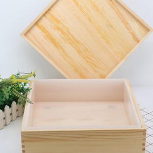 Könnte beinhalten: Eine hellfarbene Seifenform aus Holz mit abnehmbarem Deckel. Der Innenraum hat eine hellrosa Silikonauskleidung. Die Holzkiste ist rechteckig mit sichtbarer Holzmaserung. Der Deckel ist quadratisch.