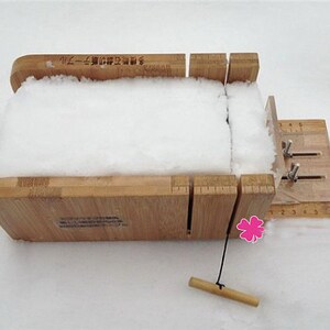 May include: A wooden soap mold with a white bar of soap inside. The mold has a ruler on the side and a lever to adjust the size of the soap. The mold is sitting on a white surface.