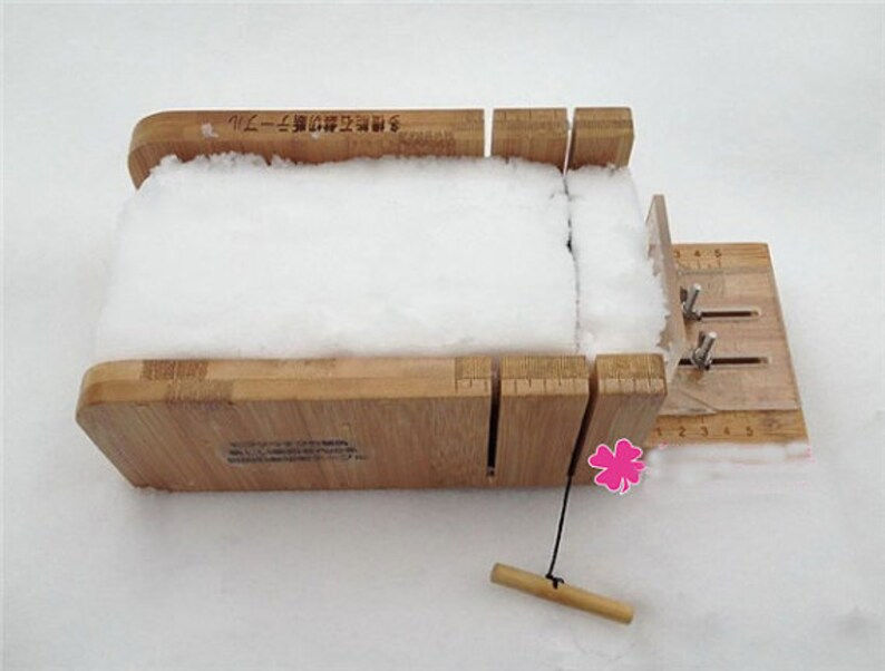 May include: A wooden soap mold with a white bar of soap inside. The mold has a ruler on the side and a lever to adjust the size of the soap. The mold is sitting on a white surface.