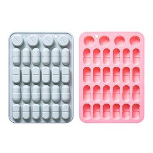 Può includere: Due stampi rettangolari in silicone, uno azzurro e uno rosa chiaro, ciascuno con una griglia di cavità a forma di compresse circolari e capsule ovali allungate. Ogni stampo contiene quattro file di capsule e una fila superiore di compresse.