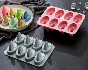 May include: A collection of silicone molds for crafting soaps or desserts. The image shows a variety of molds, including a pink oval-shaped mold, a gray tray with cone-shaped molds, and a plate with colorful cone-shaped items. The items are arranged on a dark surface.