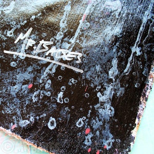 May include: Close-up of a black canvas with white paint splatters and the signature "Mr. Isaksen" in white paint.
