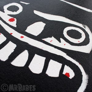 May include: Black canvas tote bag with a white graphic of a smiling face with large teeth and eyes. The graphic has red paint splatters.