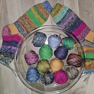 Può includere: Una ciotola di vetro piena di gomitoli di lana colorati, con due calzini a righe colorate e ferri da maglia su una superficie di legno.