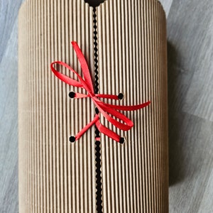 May include: A brown cardboard box with a red ribbon tied around it. The box has a corrugated texture and is tied with a bow.