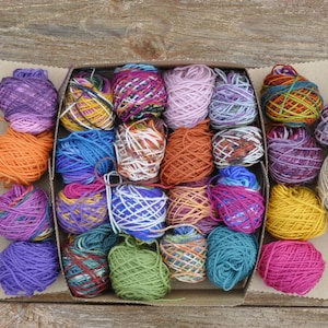 May include: A box filled with colorful yarn balls in various shades of purple, blue, green, red, orange, and yellow. The yarn is arranged in a grid pattern.
