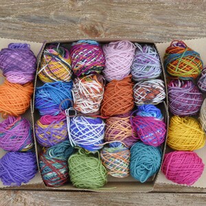 May include: A box filled with colorful yarn balls in various shades of purple, blue, green, red, orange, and yellow. The yarn is arranged in a grid pattern.