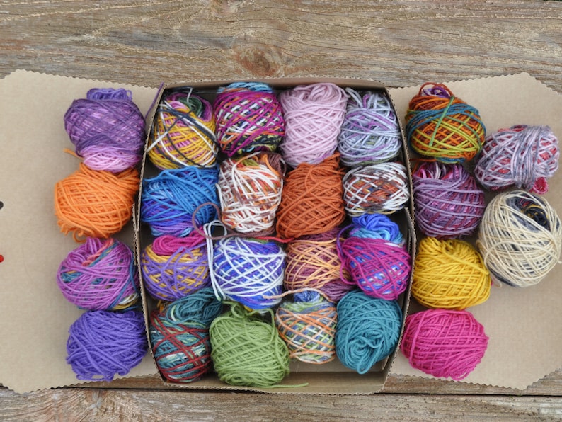 May include: A box filled with colorful yarn balls in various shades of purple, blue, green, red, orange, and yellow. The yarn is arranged in a grid pattern.