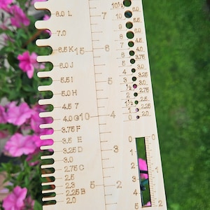 May include: A wooden knitting needle gauge with measurements in US sizes, millimeters, and centimeters. The gauge has a range of sizes from 2.0 mm to 10.0 mm.