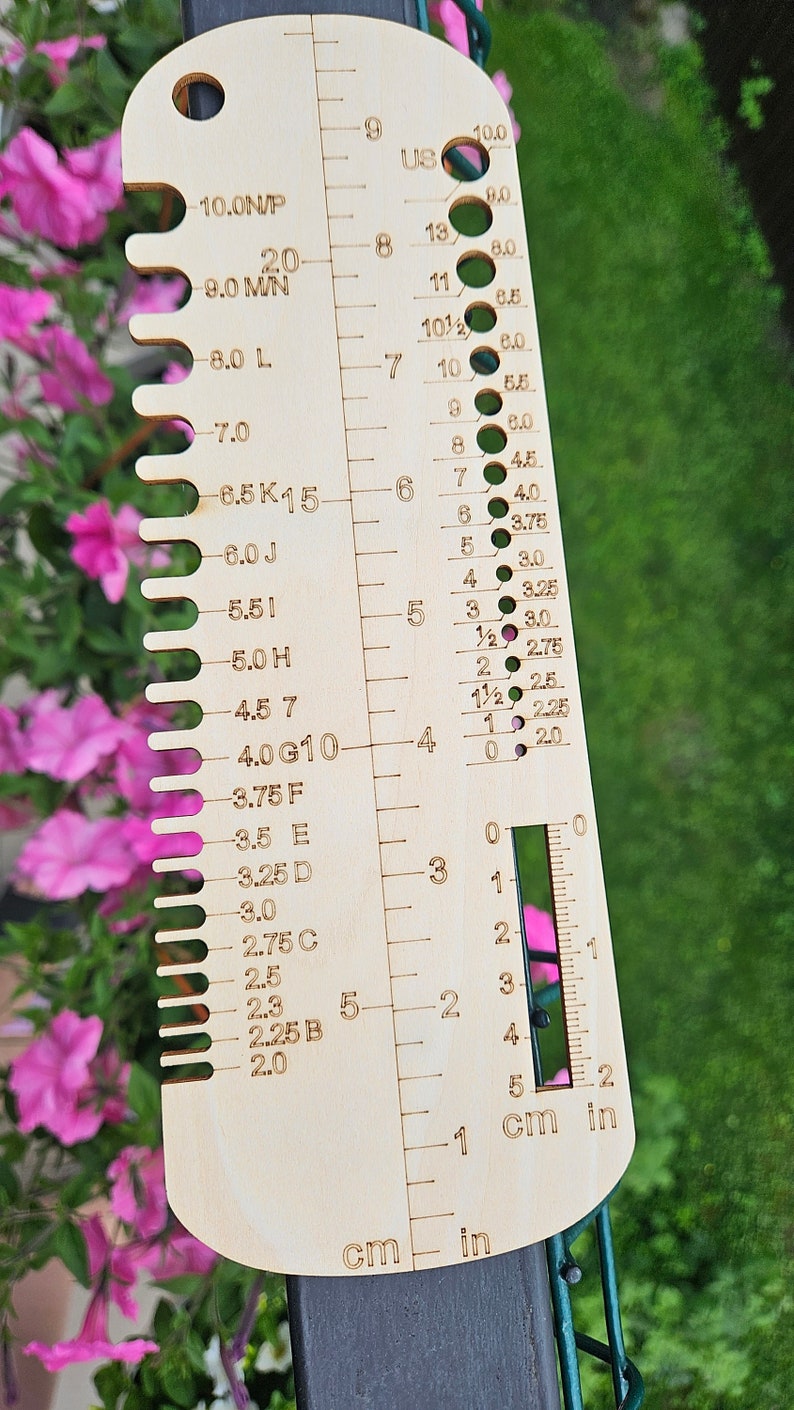May include: A wooden knitting needle gauge with measurements in US sizes, millimeters, and centimeters. The gauge has a range of sizes from 2.0 mm to 10.0 mm.
