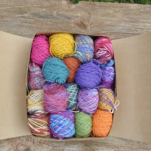 Puede incluir: Una caja de cartón llena de ovillos de lana de colores. La lana está en varios tonos de rosa, morado, azul, verde, amarillo y naranja.
