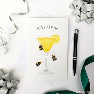 May include: A greeting card with a yellow cocktail glass with a lemon wedge and three bumblebees. The text on the card reads "Let's get buzzed".