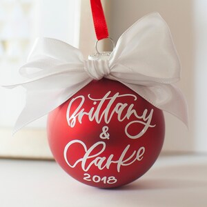 May include: Red Christmas ornament with a white bow and the text "Brittany & Clarke 2018" written in white cursive.