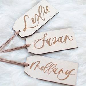 Puede incluir: Tres etiquetas de regalo de madera con cord&oacute;n de cuero marr&oacute;n. Las etiquetas est&aacute;n grabadas con los nombres "Leslie", "Susan" y "Mellary" en una fuente cursiva.