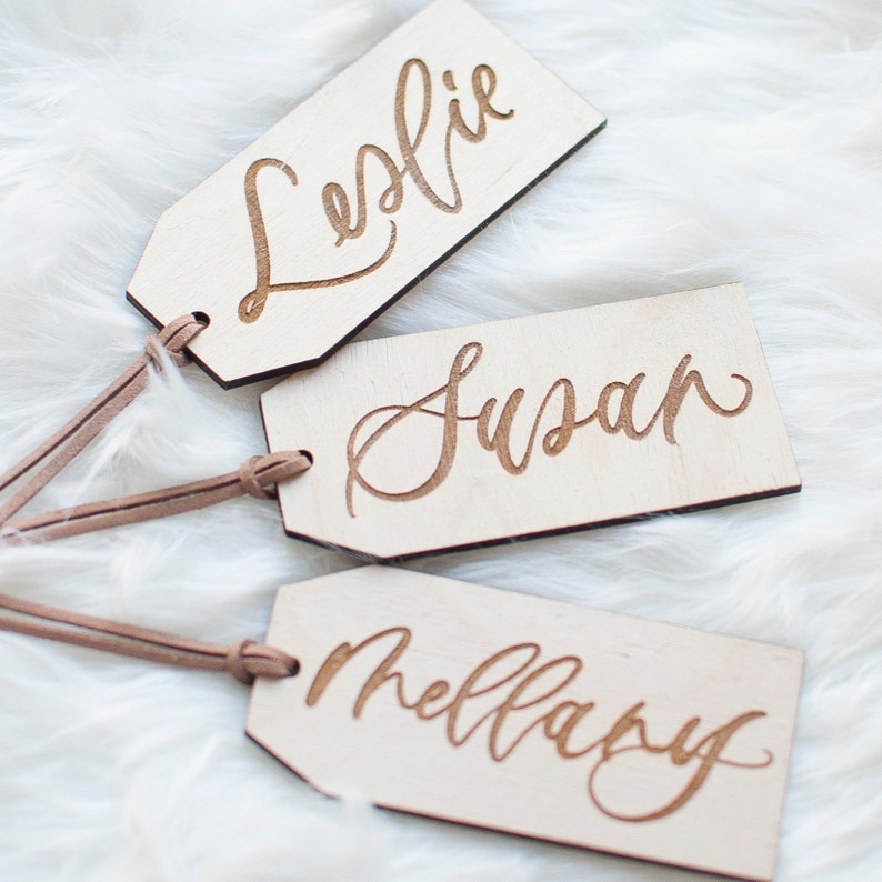 Puede incluir: Tres etiquetas de regalo de madera con cord&oacute;n de cuero marr&oacute;n. Las etiquetas est&aacute;n grabadas con los nombres "Leslie", "Susan" y "Mellary" en una fuente cursiva.