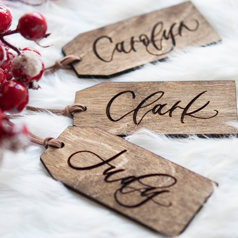 Puede incluir: Tres etiquetas de regalo de madera con los nombres "Carolyn", "Clark" y "Judy" grabados en escritura cursiva. Las etiquetas est&aacute;n atadas con un cordel marr&oacute;n y est&aacute;n sobre una superficie blanca y esponjosa.