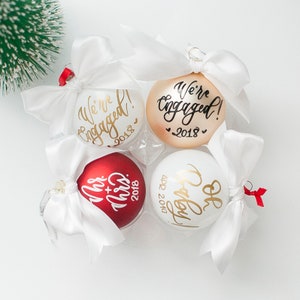 May include: Four Christmas ornaments with white satin bows. The ornaments are white, gold, and red. The white ornaments have gold lettering that says "We're Engaged!" and "Oh Happy Day". The gold ornament has black lettering that says "We're Engaged!" The red ornament has gold lettering that says "Mr. & Mrs. 2018".