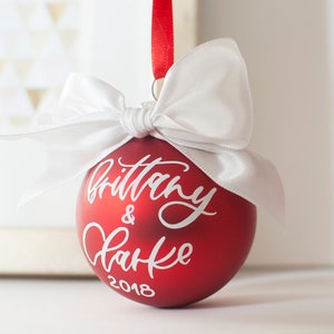 May include: Red Christmas ornament with a white bow and the names "Brittany & Clarke" written in white script with the year "2018" below.