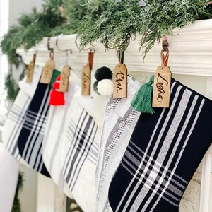 Puede incluir: Cinco calcetines de Navidad de cuadros en blanco y negro con etiquetas de madera. Los nombres en las etiquetas son "Ella", "Liam", "Liam", "Owen" y "Leila".