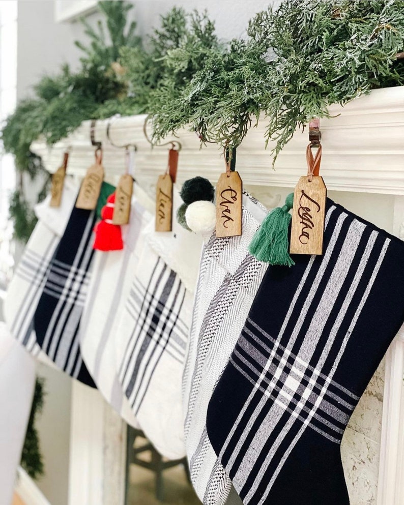 Puede incluir: Cinco calcetines de Navidad de cuadros en blanco y negro con etiquetas de madera. Los nombres en las etiquetas son "Ella", "Liam", "Liam", "Owen" y "Leila".