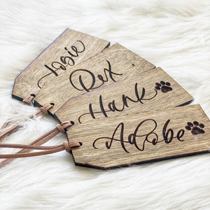 Puede incluir: Cuatro etiquetas de identificaci&oacute;n para mascotas de madera con correas de cuero. Las etiquetas est&aacute;n grabadas con los nombres "Rosie", "Rex", "Hank" y "Adobe" con un dise&ntilde;o de huella de pata.