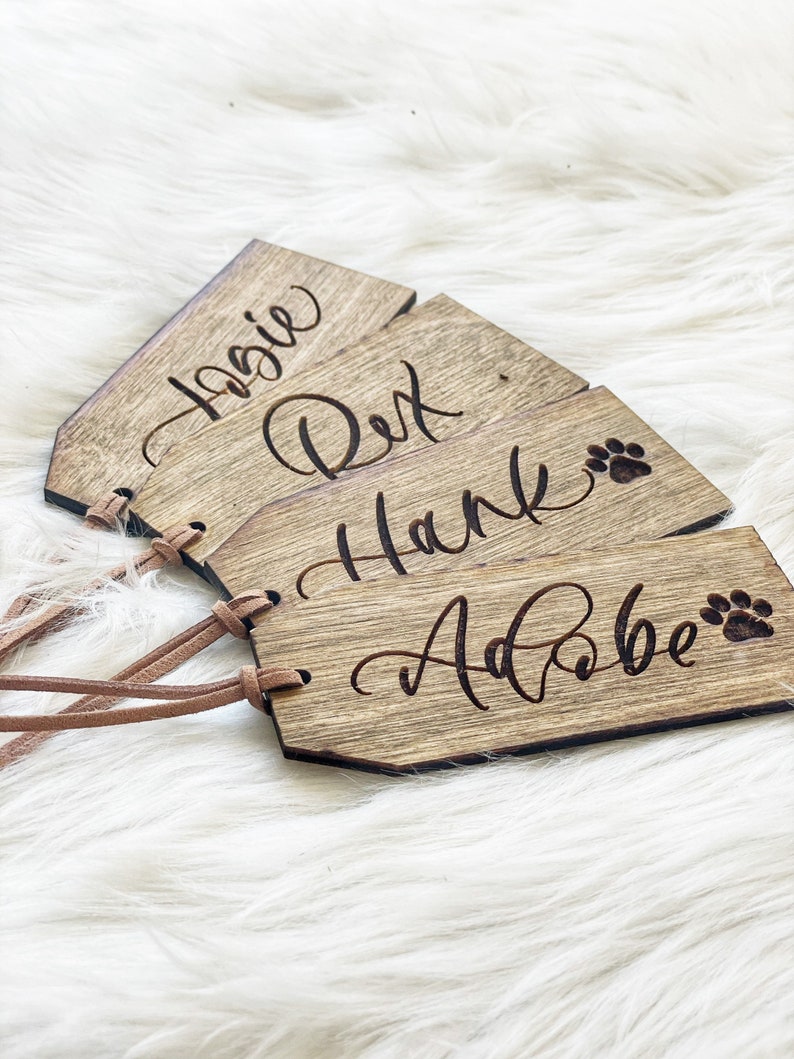 Puede incluir: Cuatro etiquetas de identificaci&oacute;n para mascotas de madera con correas de cuero. Las etiquetas est&aacute;n grabadas con los nombres "Rosie", "Rex", "Hank" y "Adobe" con un dise&ntilde;o de huella de pata.