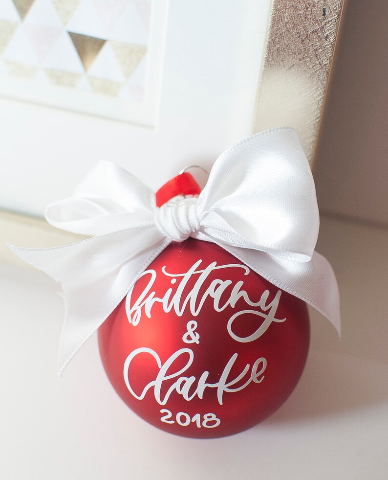 May include: Red Christmas ornament with a white bow and the text "Brittany & Clarke 2018" written in white calligraphy.