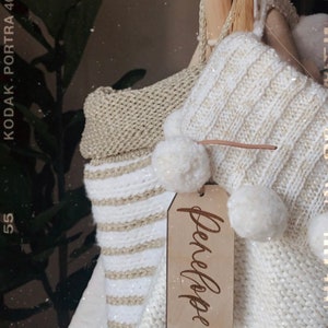 Puede incluir: Un tipi de punto de rayas blancas y doradas con pompones blancos y una etiqueta de madera que dice "Penelope".