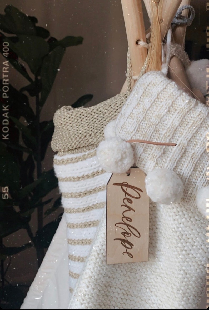Puede incluir: Un tipi de punto de rayas blancas y doradas con pompones blancos y una etiqueta de madera que dice "Penelope".