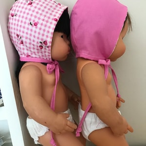May include: Two dolls wearing bonnets. One is plain pink, the other has a white and pink gingham pattern with strawberries. Both dolls wear white nappies and have pink bonnet ties.