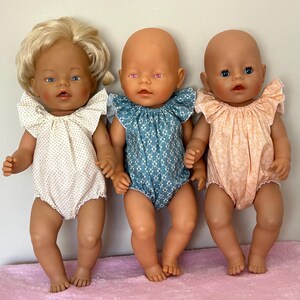 May include: Three baby dolls dressed in patterned rompers. The doll on the left has blonde hair and a white romper with small brown polka dots. The middle doll wears a blue romper with a white floral pattern. The doll on the right has a peach romper.