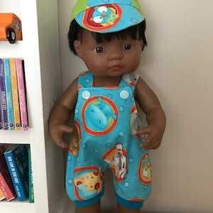 May include: A doll wearing a blue and green cap and matching dungarees. The dungarees feature a pattern of cartoon animals in orange and blue circles. The doll has dark hair and skin, and is standing in front of a bookshelf.