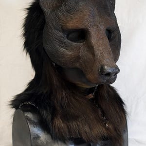 Könnte beinhalten: Eine braune Bärenmaske mit texturierter, realistischer Schnauze und Ohren, akzentuiert durch langes, tiefschwarzes Kunstfell um Hals und Kopf. Die Maske ist auf einem schwarzen, rechteckigen Ständer präsentiert und zeigt detaillierte Merkmale.