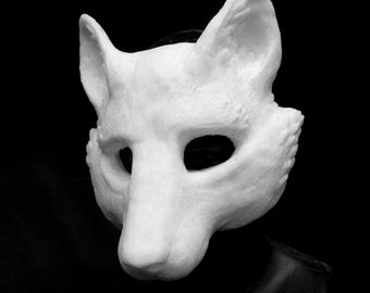 May include: A white, sculpted animal mask featuring pointed ears, a snout, and eye openings. The mask has a textured surface and is displayed on a dark, neutral form. The design covers the face and sides of the head with a smooth, matte finish.