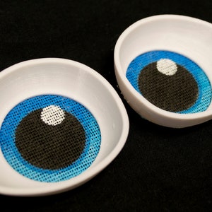 Puede incluir: Dos ojos de muñeca de plástico blanco con iris azules y pupilas negras. Los iris tienen un patrón de tejido texturizado.