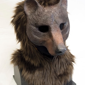 May include: A brown bear mask featuring a textured, moulded face with deep-set eye openings and a realistic snout. The mask is surrounded by thick, long, dark brown faux fur that covers the head and neck area, creating a full, shaggy appearance.