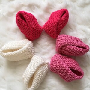 Puede incluir: Seis patucos de beb&eacute; tejidos a mano en tonos rojos, rosas y blancos est&aacute;n dispuestos sobre una superficie blanca y esponjosa. Los patucos est&aacute;n hechos de hilo suave y tienen un dise&ntilde;o sencillo y cl&aacute;sico.