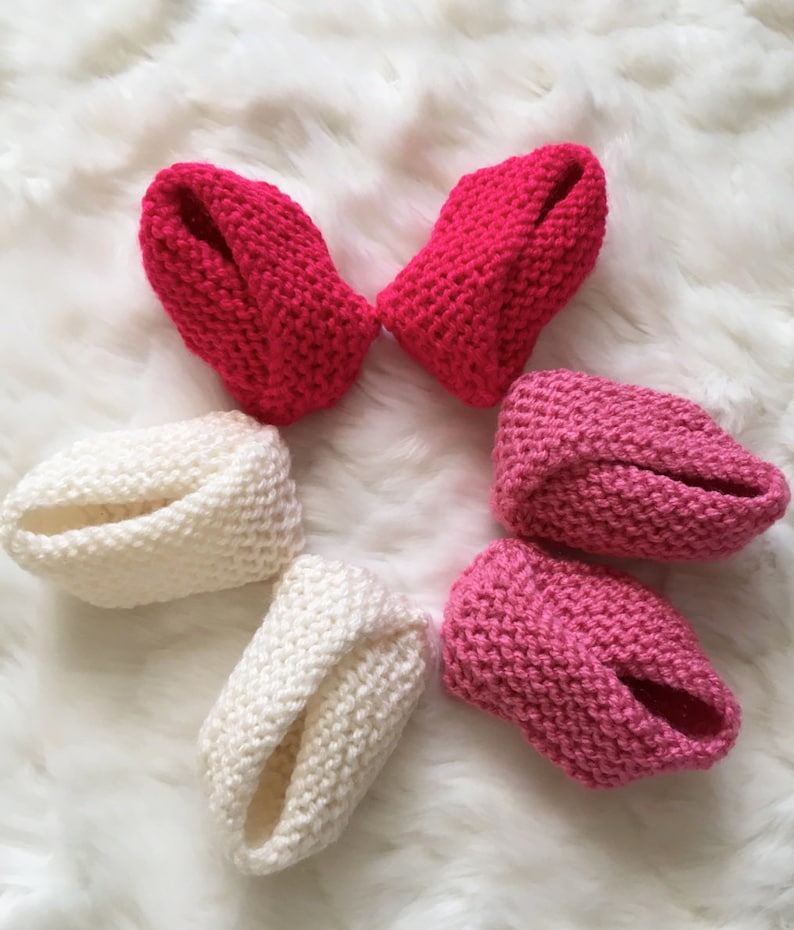 Puede incluir: Seis patucos de beb&eacute; tejidos a mano en tonos rojos, rosas y blancos est&aacute;n dispuestos sobre una superficie blanca y esponjosa. Los patucos est&aacute;n hechos de hilo suave y tienen un dise&ntilde;o sencillo y cl&aacute;sico.